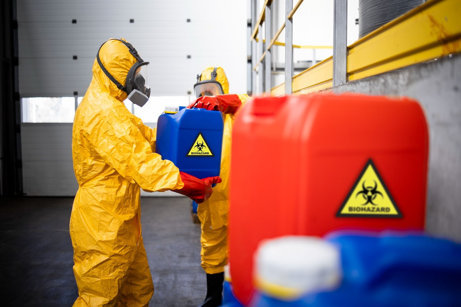 Trabalhadores usando trajes de proteção e máscaras manuseando recipientes identificados com símbolo de biohazard, representando riscos químicos em ambiente controlado.