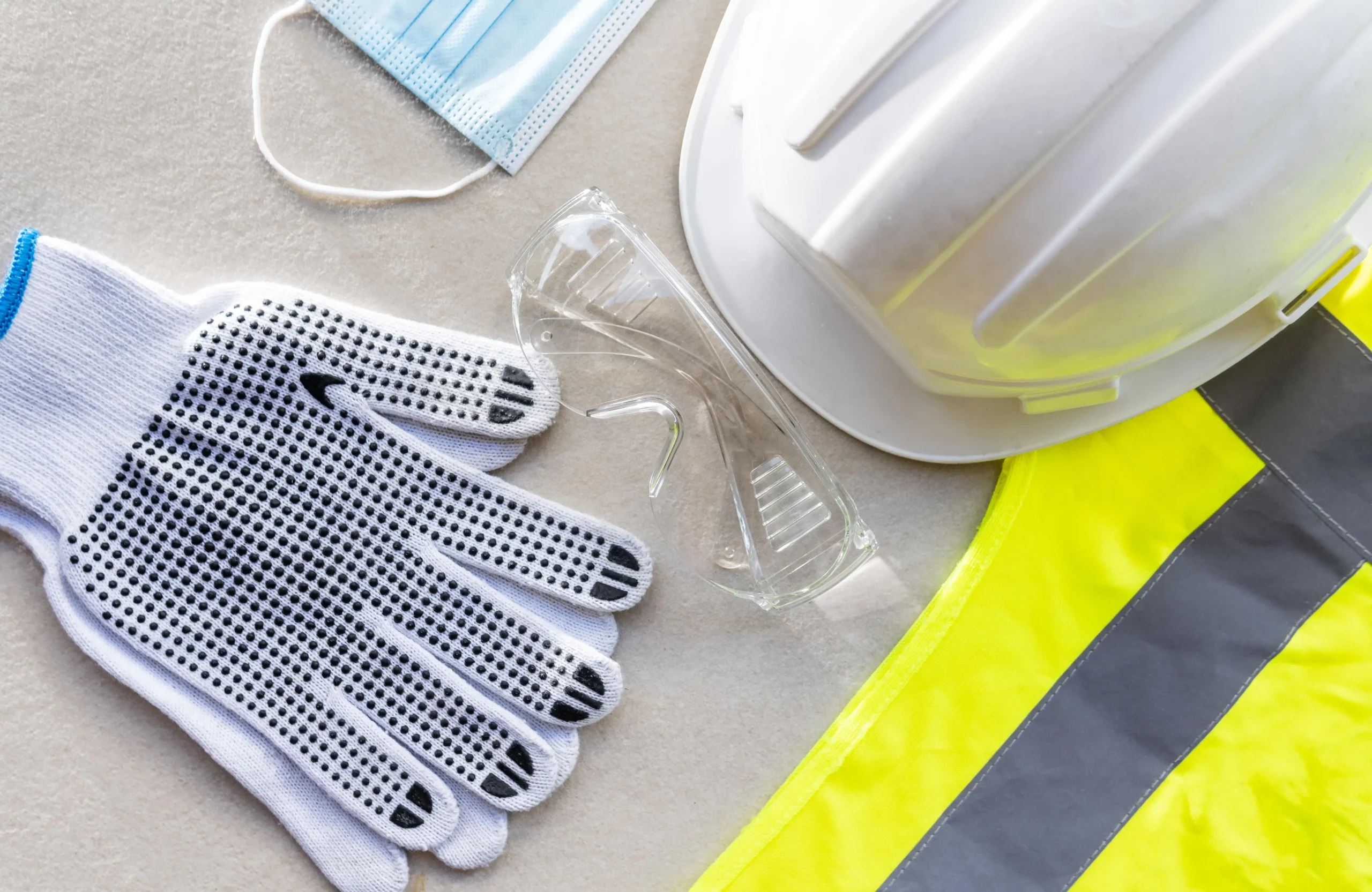 Capacete de segurança, luvas e máscara vistos de cima, ilustrando equipamentos de proteção individual exemplos aplicados em ambientes de trabalho.